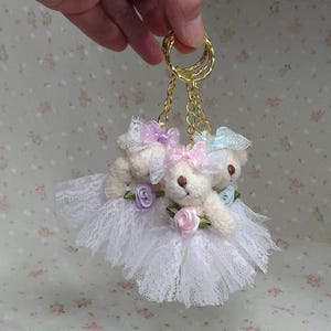 May include: A white lace keychain with three small teddy bears wearing pastel colored bows and flowers. The keychain has a gold chain and ring.