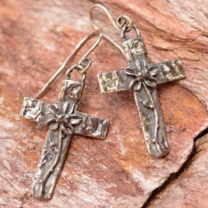 May include: A pair of silver cross earrings with a textured finish. Each cross features a floral design at the center and along the vertical arm. The earrings hang from simple silver hooks, showcasing a rustic, handcrafted aesthetic.