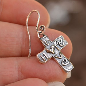 May include: Silver cross dangle earring with the word "love" inscribed on it. The cross is textured and has a heart shape in the center.