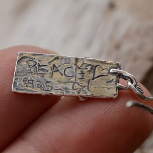 May include: A silver earring with a rectangular pendant. The pendant has the word "PEACE" and the numbers "1925" etched into it, along with a heart symbol. The earring is attached to a silver hook.