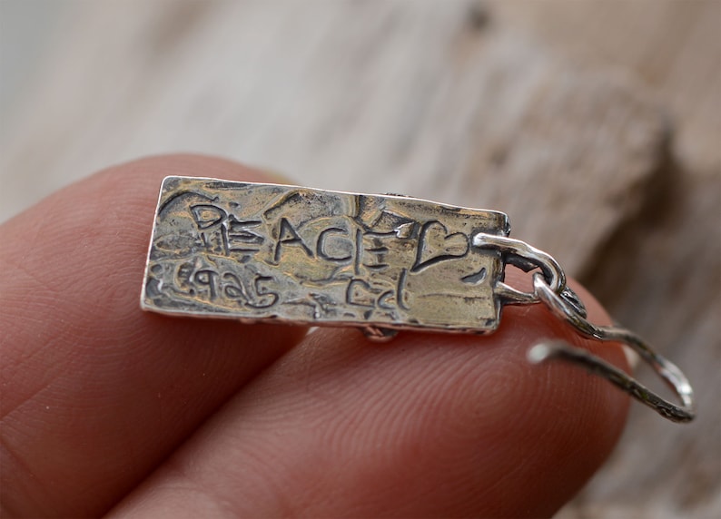 May include: A silver earring with a rectangular pendant. The pendant has the word "PEACE" and the numbers "1925" etched into it, along with a heart symbol. The earring is attached to a silver hook.