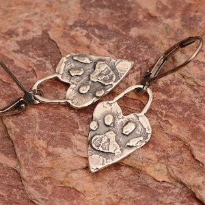 May include: A pair of silver-toned drop earrings featuring heart-shaped pendants with a textured, oxidized finish. Each heart displays a raised paw print design in the center. The earrings are attached to silver-toned lever-back ear wires.