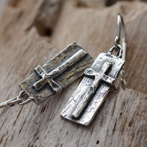 May include: A pair of silver earrings featuring a cross design. Each earring has a rectangular base with a textured surface and a detailed cross. The earrings are displayed on a wooden surface.