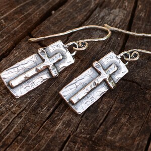 May include: A pair of silver earrings featuring a cross design. Each earring has a rectangular base with a textured surface and a raised cross. The earrings have a hook closure. The background is a wooden surface.