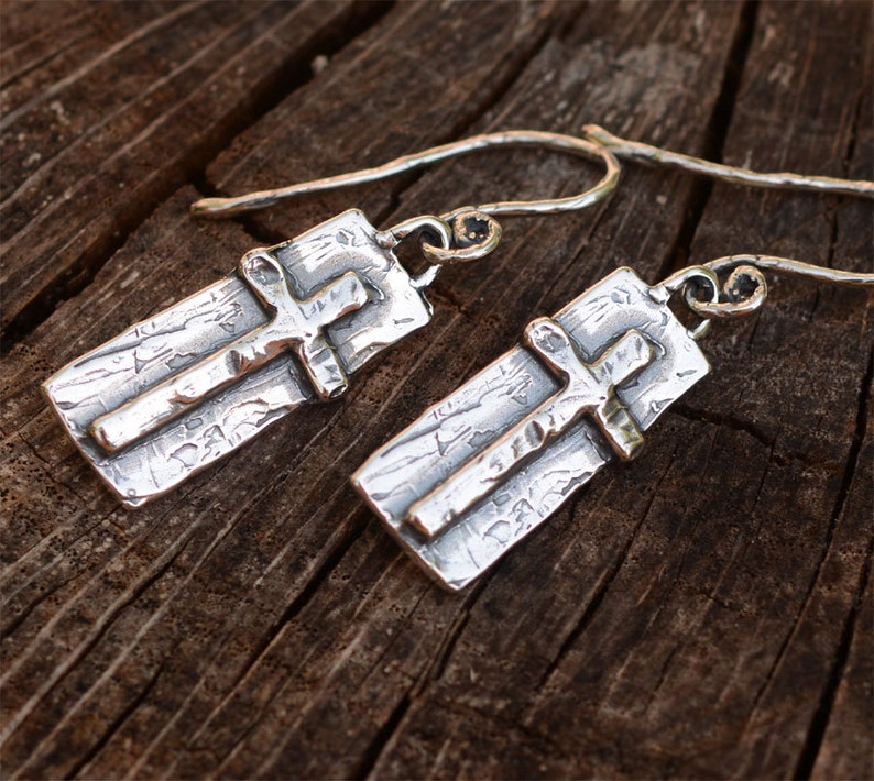 May include: A pair of silver earrings featuring a cross design. Each earring has a rectangular base with a textured surface and a raised cross. The earrings have a hook closure. The background is a wooden surface.