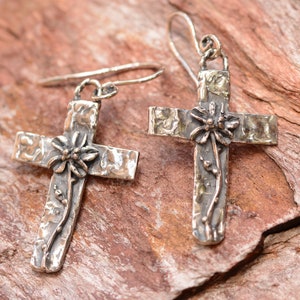 May include: Silver-toned cross earrings with a textured finish. Each cross features a floral design at the center and a vine-like accent. The earrings have a hook closure and are displayed on a reddish-brown surface.