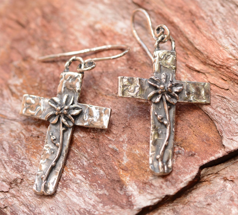 May include: Silver-toned cross earrings with a textured finish. Each cross features a floral design at the center and a vine-like accent. The earrings have a hook closure and are displayed on a reddish-brown surface.