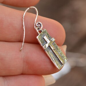 May include: A silver earring with a rectangular pendant featuring a cross design. The earring has a textured surface and a curved hook for the ear. The cross is raised and has a contrasting finish.