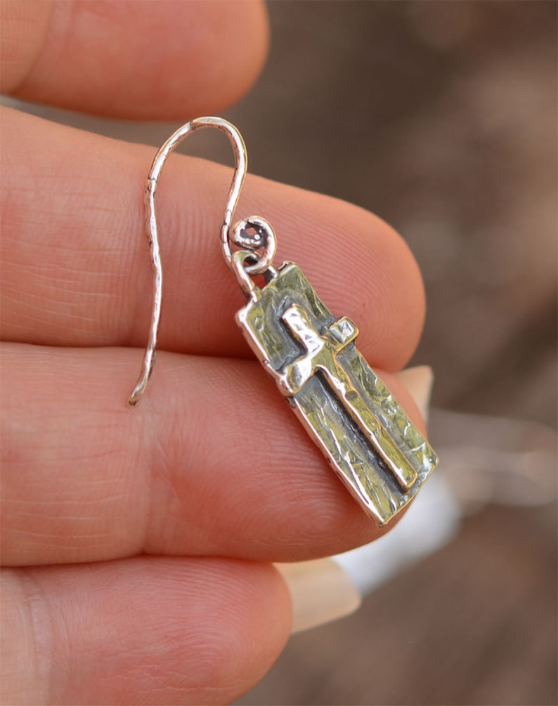 May include: A silver earring with a rectangular pendant featuring a cross design. The earring has a textured surface and a curved hook for the ear. The cross is raised and has a contrasting finish.