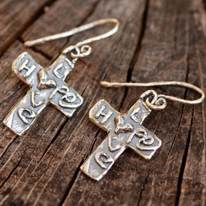 May include: A pair of silver cross earrings with a hammered texture and the word "Hope" inscribed on each cross.