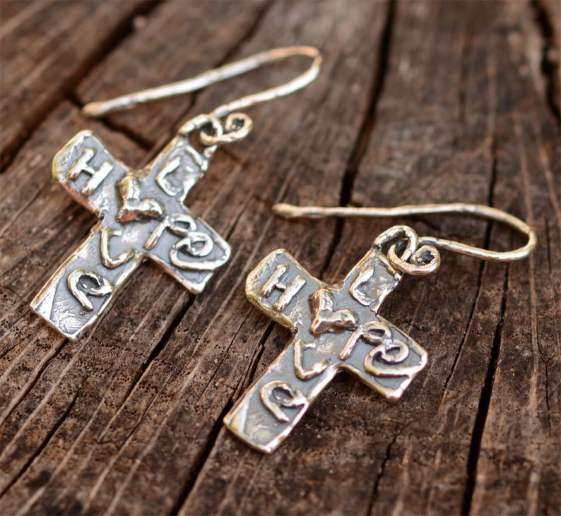 May include: A pair of silver cross earrings with a hammered texture and the word "Hope" inscribed on each cross.
