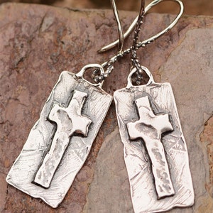 May include: A pair of silver-toned earrings featuring a rectangular pendant with a textured surface and a raised cross design. The earrings have a hook closure and are displayed on a stone surface.