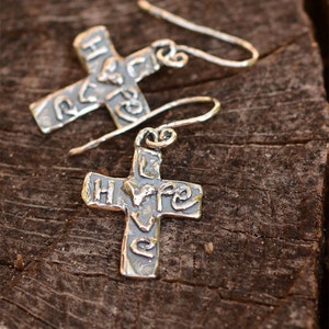 May include: Silver cross earrings with the word "love" embossed on the front. The earrings are hanging from a wooden surface.