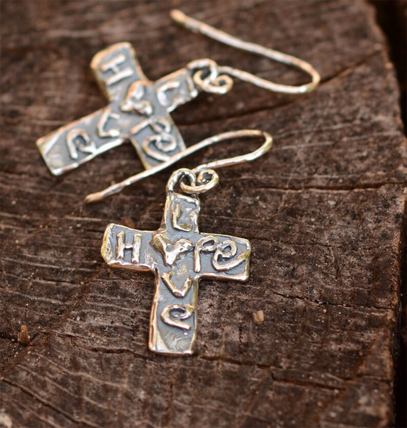 May include: Silver cross earrings with the word "love" embossed on the front. The earrings are hanging from a wooden surface.
