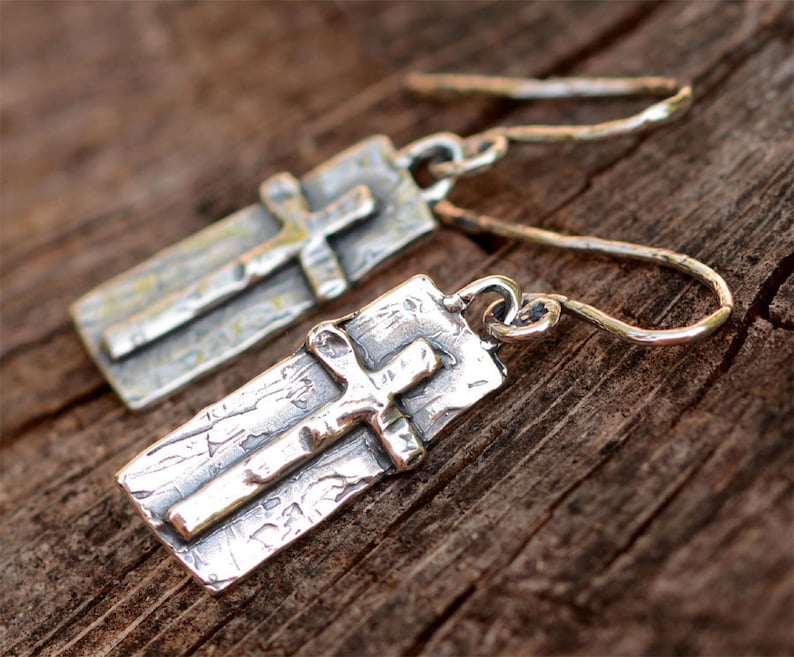 May include: A pair of silver-toned earrings featuring a cross design. Each earring has a rectangular base with a textured surface and a smaller cross overlay. The earrings have a hook closure.