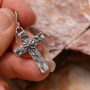 May include: A silver cross earring with a textured surface and floral detailing. The cross features a flower at the center and vines along the arms. The earring is held by a silver hook.