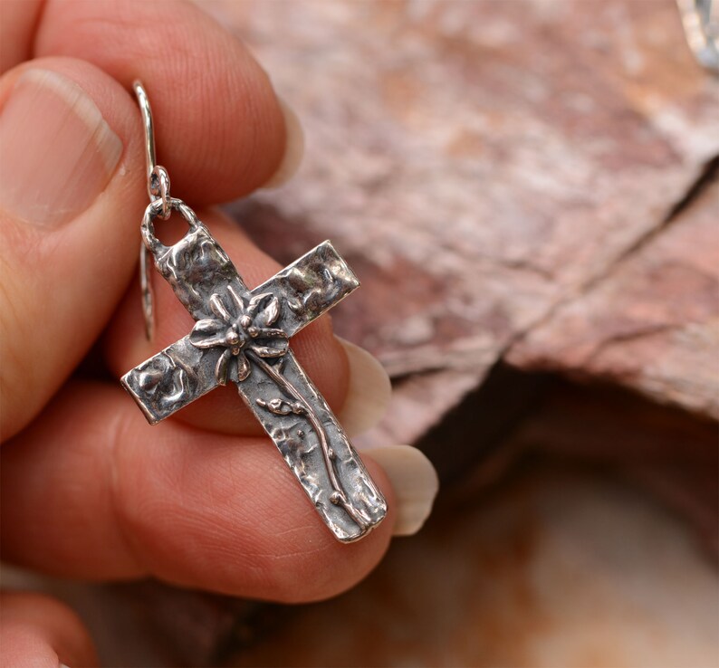 May include: A silver cross earring with a textured surface and floral detailing. The cross features a flower at the center and vines along the arms. The earring is held by a silver hook.