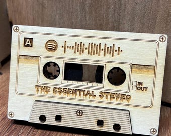 May include: A rectangular light-colored wooden plaque shaped like a cassette tape. It features a Spotify code, the text "THE ESSENTIAL STEVES," a central window, two circular spools, and four small screw-like details at the corners.