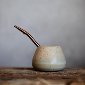 Peut inclure: Une calebasse à maté en céramique blanc cassé moucheté avec une paille en métal couleur cuivre. La calebasse a une base arrondie et un bord légèrement évasé. La paille est insérée en biais.