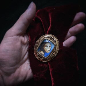 May include: A gold-toned brooch with a detailed design features a miniature portrait of a person with light brown hair and blue eyes. The portrait is set against a blue background.