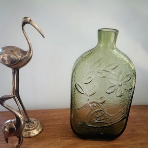 Può includere: Un vaso di vetro verde con una superficie testurizzata e un design in rilievo di un uccello e fiori. Il vaso è su una superficie di legno.