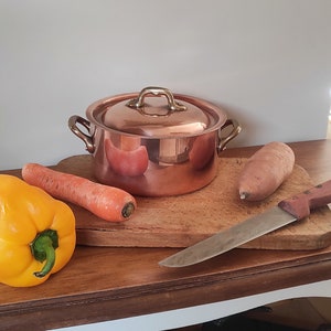 May include: A copper pot with brass handles and a lid sits on a wooden cutting board with a yellow bell pepper, a carrot, and a sweet potato. A silver knife is also on the board.