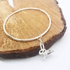 May include: A silver-colored bangle bracelet with a butterfly charm. The butterfly charm is intricately designed with cut-out details. The bracelet is displayed on a piece of wood with a light-colored background.