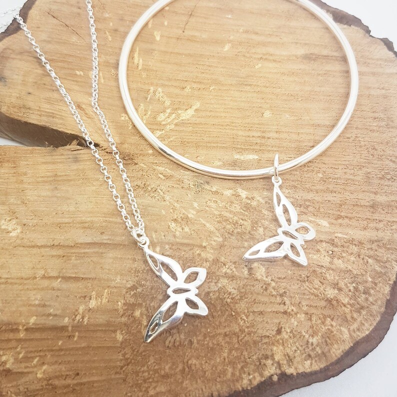 May include: A silver necklace with a butterfly pendant and a matching silver bangle bracelet. The butterfly pendant and charm have a cut-out design. The jewelry is displayed on a wooden surface.
