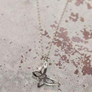 May include: A silver-colored butterfly pendant necklace. The pendant is a stylized butterfly with open wings, suspended from a delicate chain. The necklace is displayed against a mottled, neutral-toned background.