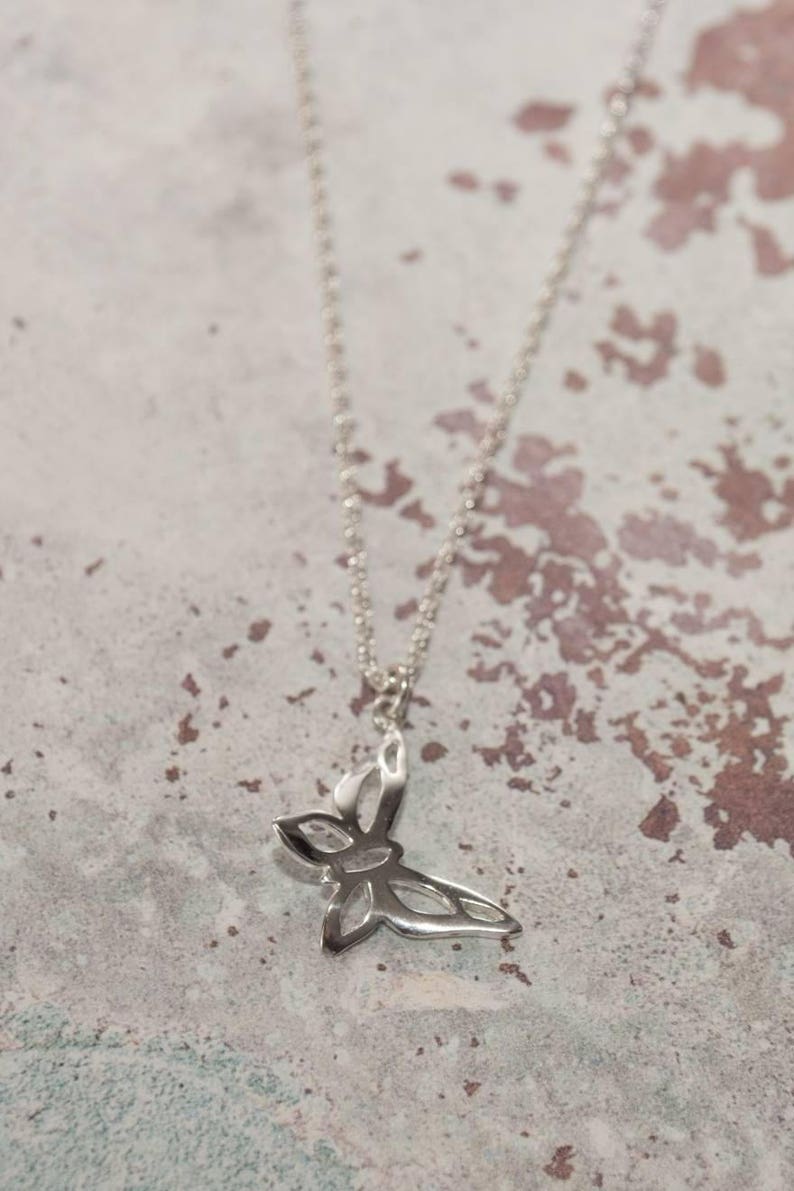 May include: A silver-colored butterfly pendant necklace. The pendant is a stylized butterfly with open wings, suspended from a delicate chain. The necklace is displayed against a mottled, neutral-toned background.