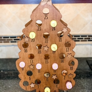 May include: Wooden Christmas tree advent calendar with 25 numbered circles. The tree is brown and has a star on top. The text "Making Spirits Bright" is engraved on the star.