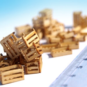 May include: A pile of small, wooden crates stacked on a white surface. The crates are made of light brown wood and have a simple, rustic design. A blue ruler is visible in the background.