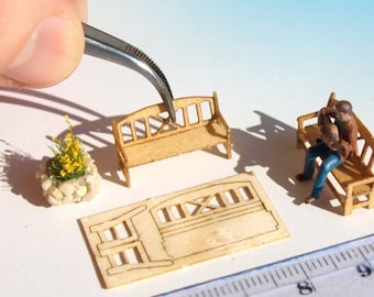 May include: A miniature wooden bench with a person sitting on it. There is a second bench and a small flower pot with yellow flowers. The benches are made of unfinished wood and are about 2 inches long. The flower pot is about 1 inch in diameter.