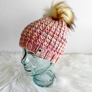May include: A pink and white knitted beanie with a brown faux fur pom pom. The beanie has a small brown leather tag with the letters "HS" on it.