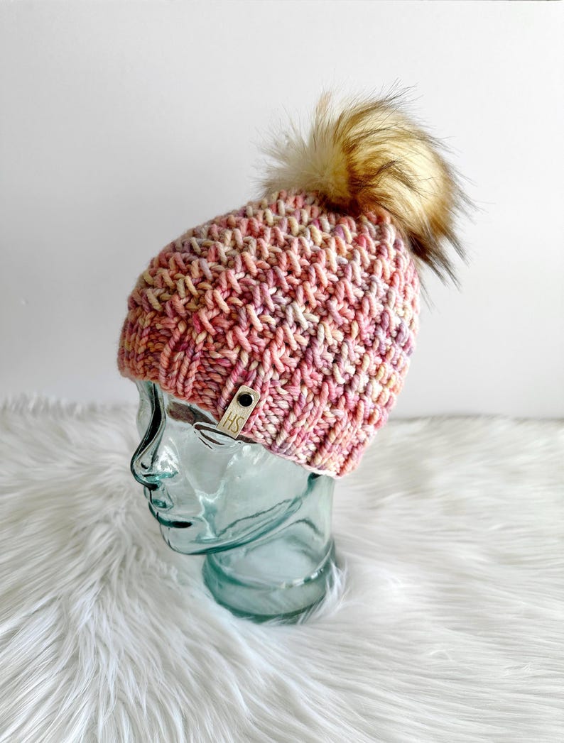 May include: A pink and white knitted beanie with a brown faux fur pom pom. The beanie has a small brown leather tag with the letters "HS" on it.