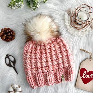 May include: A pink knitted beanie with a fluffy white pom pom. The beanie has a textured knit pattern and a small brown leather tag with the letters "HS" on it.