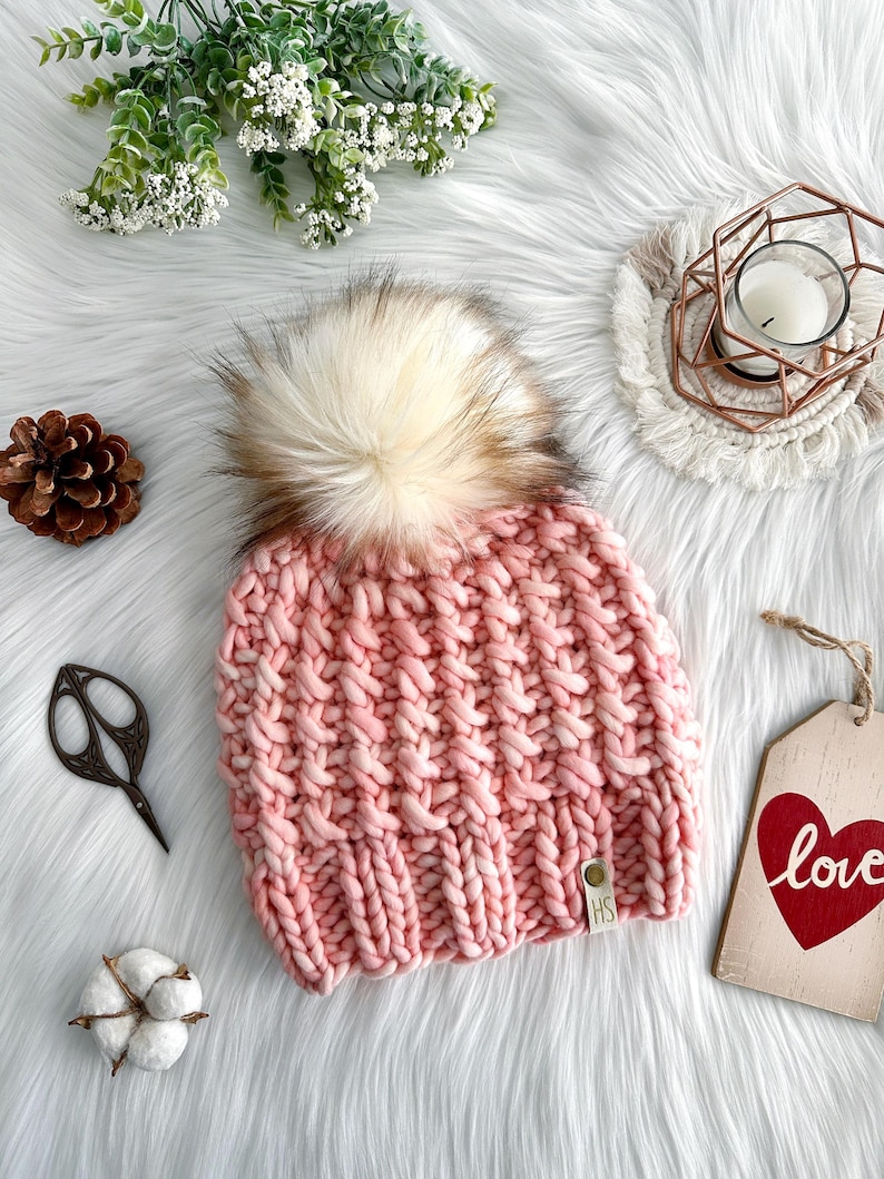May include: A pink knitted beanie with a fluffy white pom pom. The beanie has a textured knit pattern and a small brown leather tag with the letters "HS" on it.