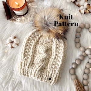 May include: A white knitted beanie with a large, fluffy pom pom made of brown, black, and white fur. The beanie has a cable knit pattern and a small leather tag with the letters "HS" on it. The beanie is on a white fluffy surface with a wooden bead garland and a lit candle in the background. The text "Knit Pattern" is written in black letters on the image.