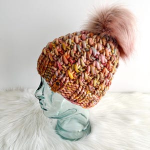 May include: A colorful knitted beanie with a fluffy pink pom pom. The beanie is made with a chunky yarn and has a unique textured stitch pattern.