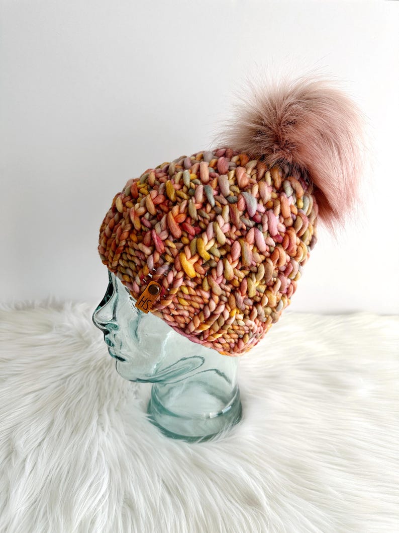 May include: A colorful knitted beanie with a fluffy pink pom pom. The beanie is made with a chunky yarn and has a unique textured stitch pattern.