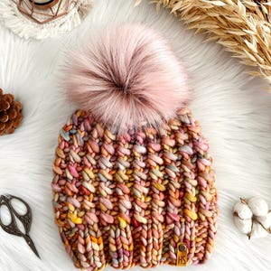 May include: A colorful knitted beanie with a fluffy pink pom pom. The beanie has a variety of colors, including pink, orange, yellow, and brown. The beanie has a leather tag with the letters "HS" on it.
