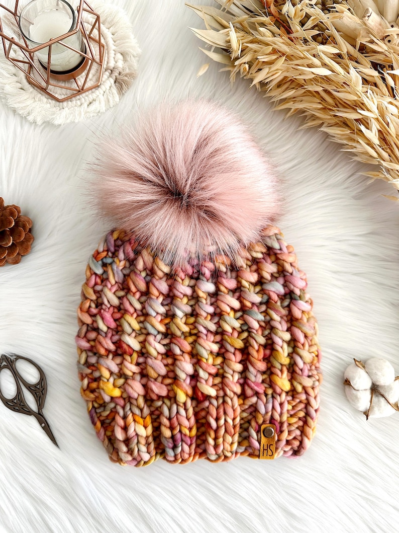 May include: A colorful knitted beanie with a fluffy pink pom pom. The beanie has a variety of colors, including pink, orange, yellow, and brown. The beanie has a leather tag with the letters "HS" on it.