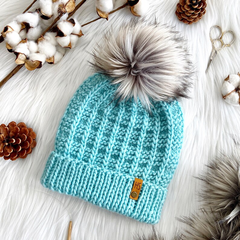 May include: A light blue knitted beanie with a faux fur pom pom. The beanie has a ribbed texture and a small leather tag with the letters "HS" on it.