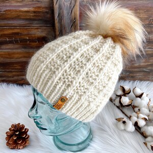 May include: A cream-colored knitted beanie with a brown faux fur pom pom. The beanie has a leather tag with the initials "HS" on it.