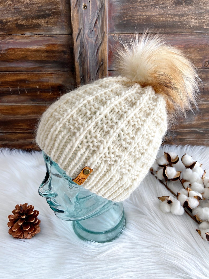 May include: A cream-colored knitted beanie with a brown faux fur pom pom. The beanie has a leather tag with the initials "HS" on it.