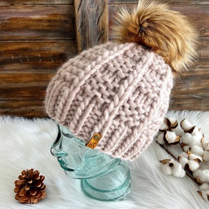 May include: A pink knitted beanie with a brown faux fur pom pom. The beanie has a leather tag with the initials "HS" on it.