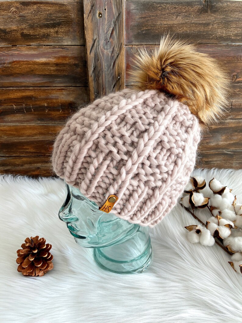 May include: A pink knitted beanie with a brown faux fur pom pom. The beanie has a leather tag with the initials "HS" on it.