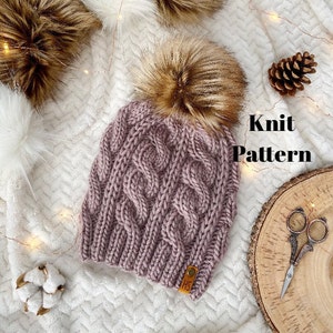 May include: A knitted hat with a cable knit pattern in a mauve colour. The hat has a brown faux fur pom pom on top. The hat is lying on a white fluffy surface with a brown leather tag that says "HS".