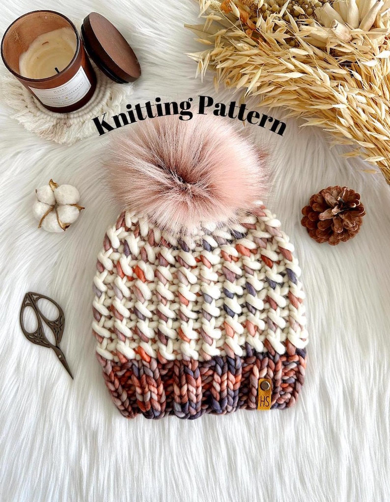 May include: Knitting pattern for a white and pink beanie with a fluffy pom pom. The beanie has a textured stitch pattern and a brown leather tag with the letters "HS" on it.
