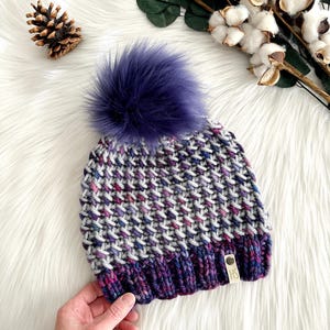 May include: A gray, purple, and pink knit beanie with a purple faux fur pom pom. The beanie has a small brown tag with the letters "HS" on it.